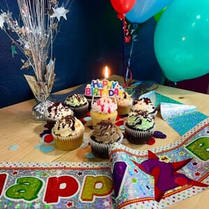 a wooden table displays six decorated cupcakes, one with a lit "happy birthday" candle. balloons, a banner, and star themed decorations enhance the festive setup. colorful confetti is spread around, creating an ambiance reminiscent of celebrations in psychology practice blue mountain nsw.