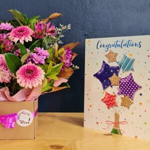 a bouquet of pink flowers in a box next to a celebratory greeting card with colorful star designs and the word "congratulations" written on it, placed on a wooden surface against a desk at Healthy Connection Clinical Psychology Blaxland